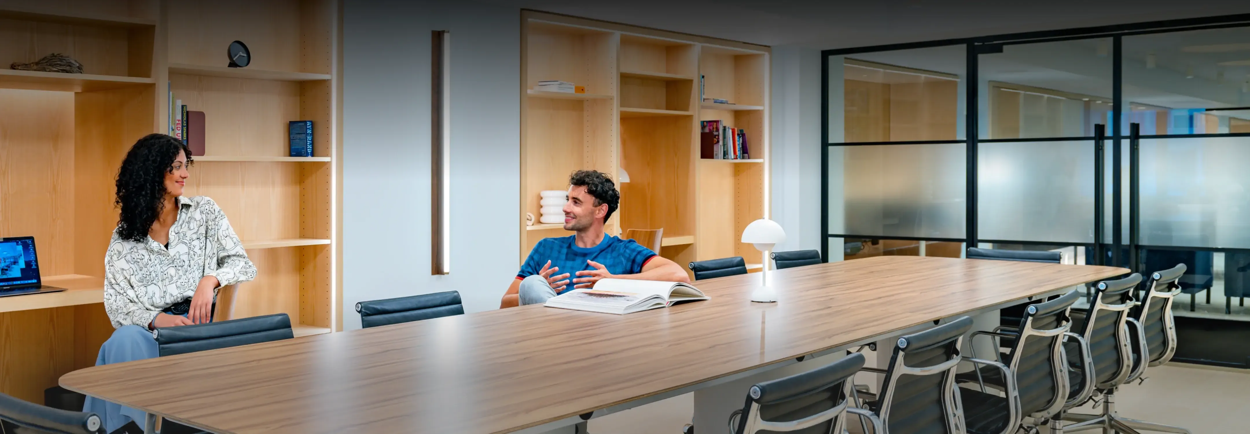 Two colleagues engaged in a casual discussion at a large conference table in the modern, well-lit meeting room of Helena 57 West.