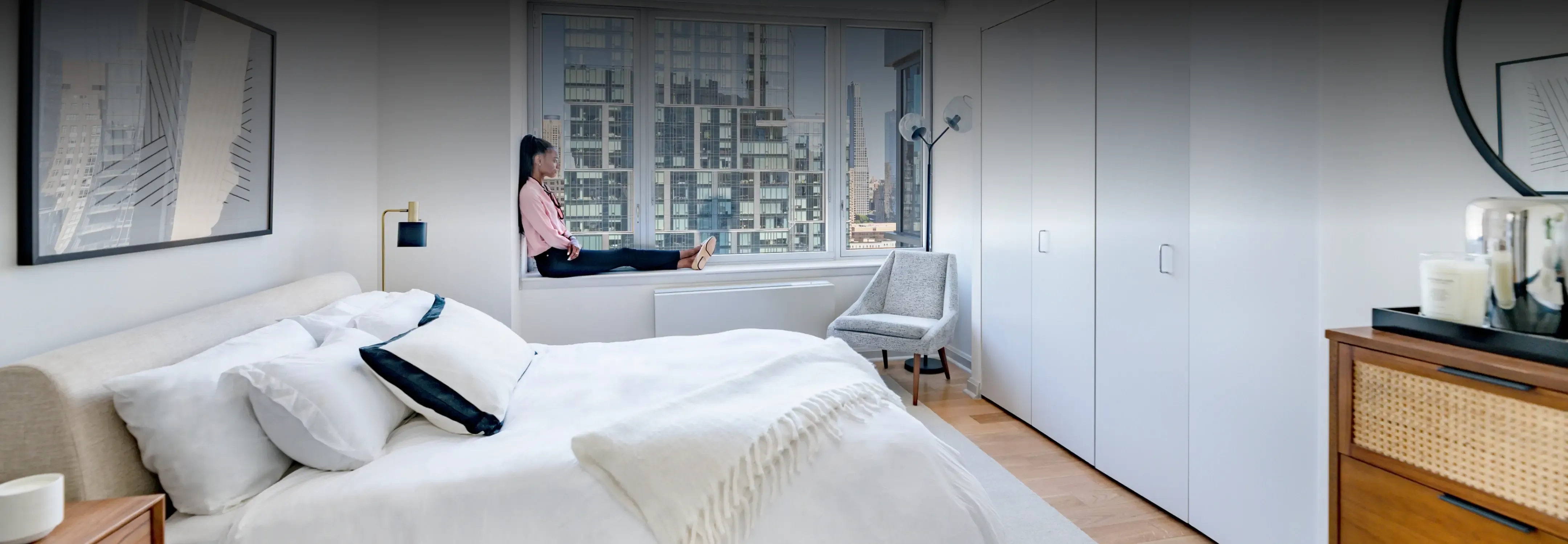 Cozy bedroom at Helena 57 West featuring a comfortable bed with cityscape views through large windows, and a person enjoying the vista.