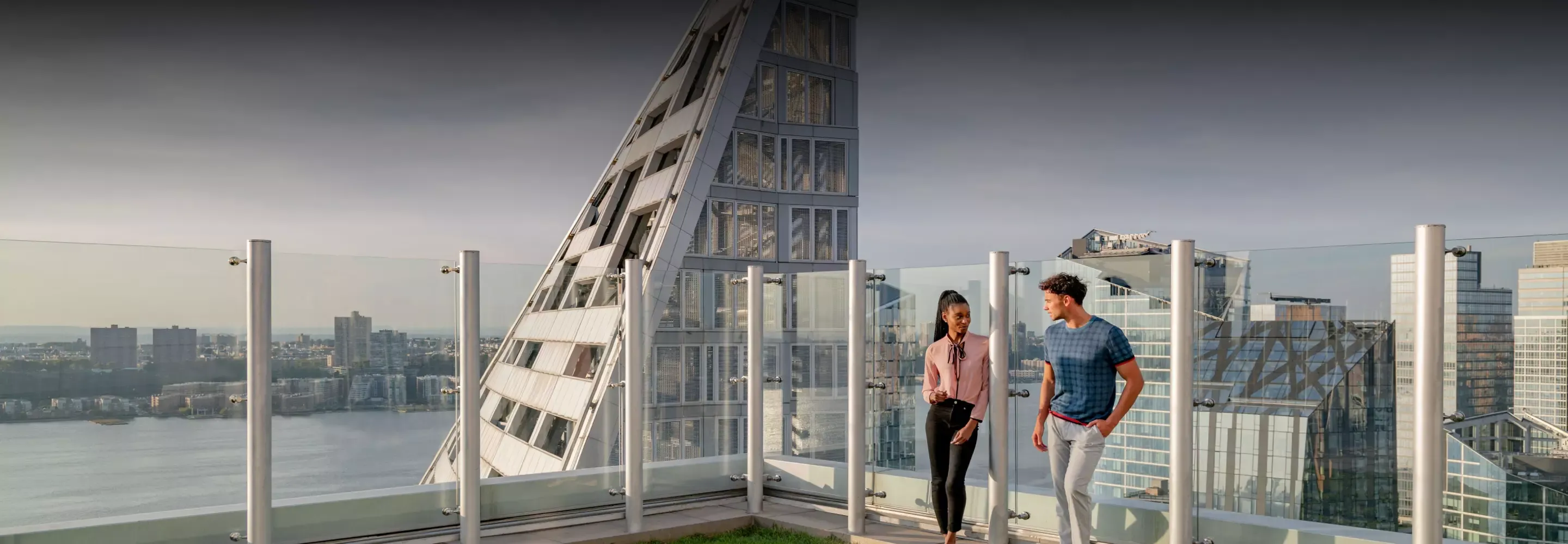 A man and woman conversing on the rooftop terrace of Helena 57 West with panoramic views of the city and water in the background.